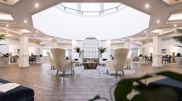 A spacious and bright senior living facility common area with a circular seating arrangement of beige armchairs around a central coffee table on a patterned rug. The room features large windows near the ceiling allowing natural light to flood in, white columns, wooden flooring, and additional seating areas with tables and chairs in the background. Green plants are placed throughout the space, enhancing the welcoming atmosphere.