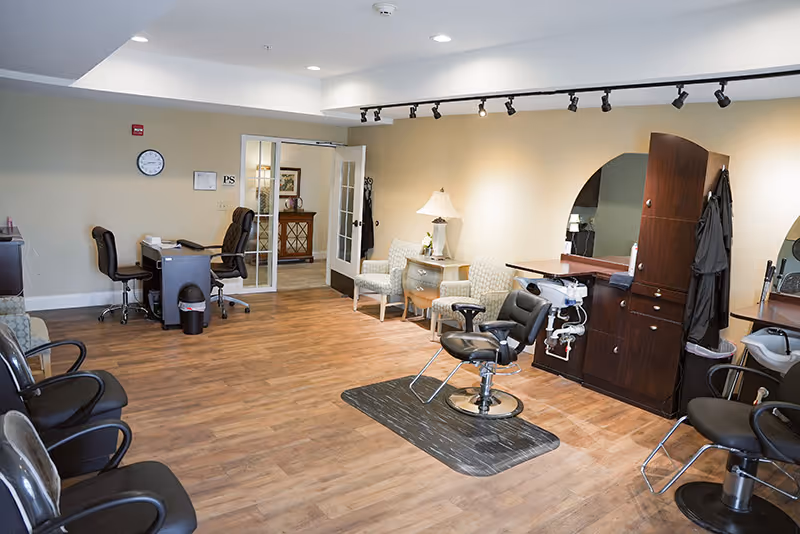 Interior view of a senior living facility's salon area with multiple black salon chairs, a styling station with mirrors, and a waiting area with cushioned chairs and a small table with a lamp. The room has wooden flooring and light-colored walls with a clock and some framed items on the wall.