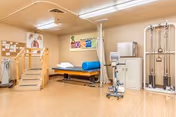 A physical therapy or rehabilitation room with wooden stairs for practice, an examination table with a blue cylindrical cushion, exercise equipment including a stationary bike and a pulley machine, and anatomical charts on the wall.