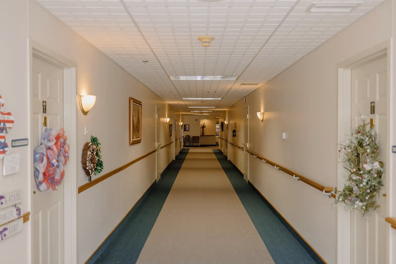 A long, well-lit hallway in an assisted living facility with beige walls, handrails on both sides, and several doors decorated with wreaths and signs. The floor has a carpet with a beige center and green edges. There are wall-mounted lights and framed artwork along the hallway, with chairs and a flower arrangement visible at the far end.