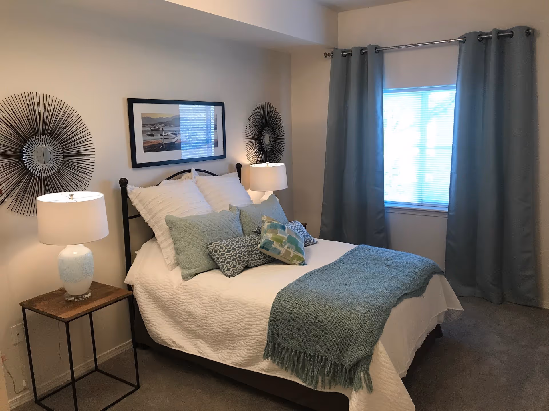 A cozy bedroom with a neatly made bed featuring white bedding and multiple decorative pillows in shades of green and blue. There are two bedside tables with white lamps on either side of the bed. The walls are decorated with two round sunburst mirrors and a framed picture above the bed. A window with gray curtains allows natural light into the room.