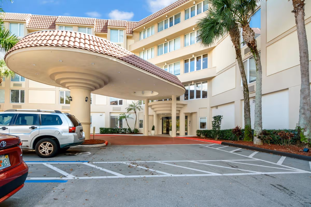Exterior view of the entrance to The Atrium at Boca Raton senior living facility, showing a covered drop-off area with a tiled roof supported by columns, parked cars, palm trees, and a multi-story beige building under a blue sky.