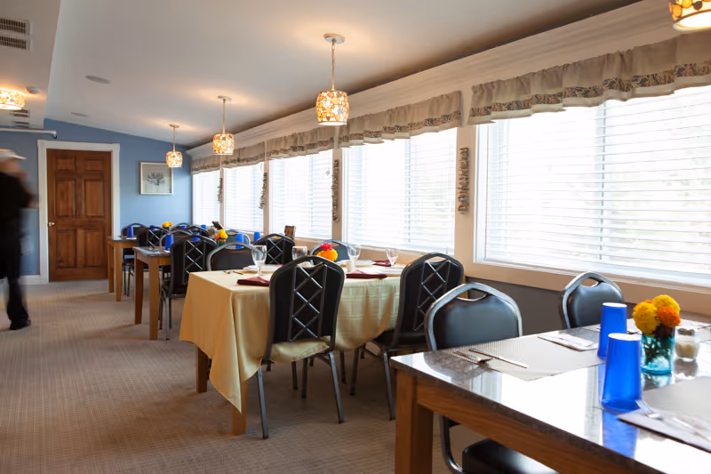 Bright dining room with tables set with tablecloths, chairs, pendant lights, and large windows with blinds.