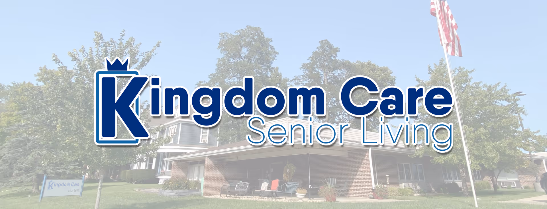 Exterior view of Kingdom Care Senior Living facility showing a brick building with a covered entrance, surrounded by trees and a lawn, with an American flag on a flagpole to the right.