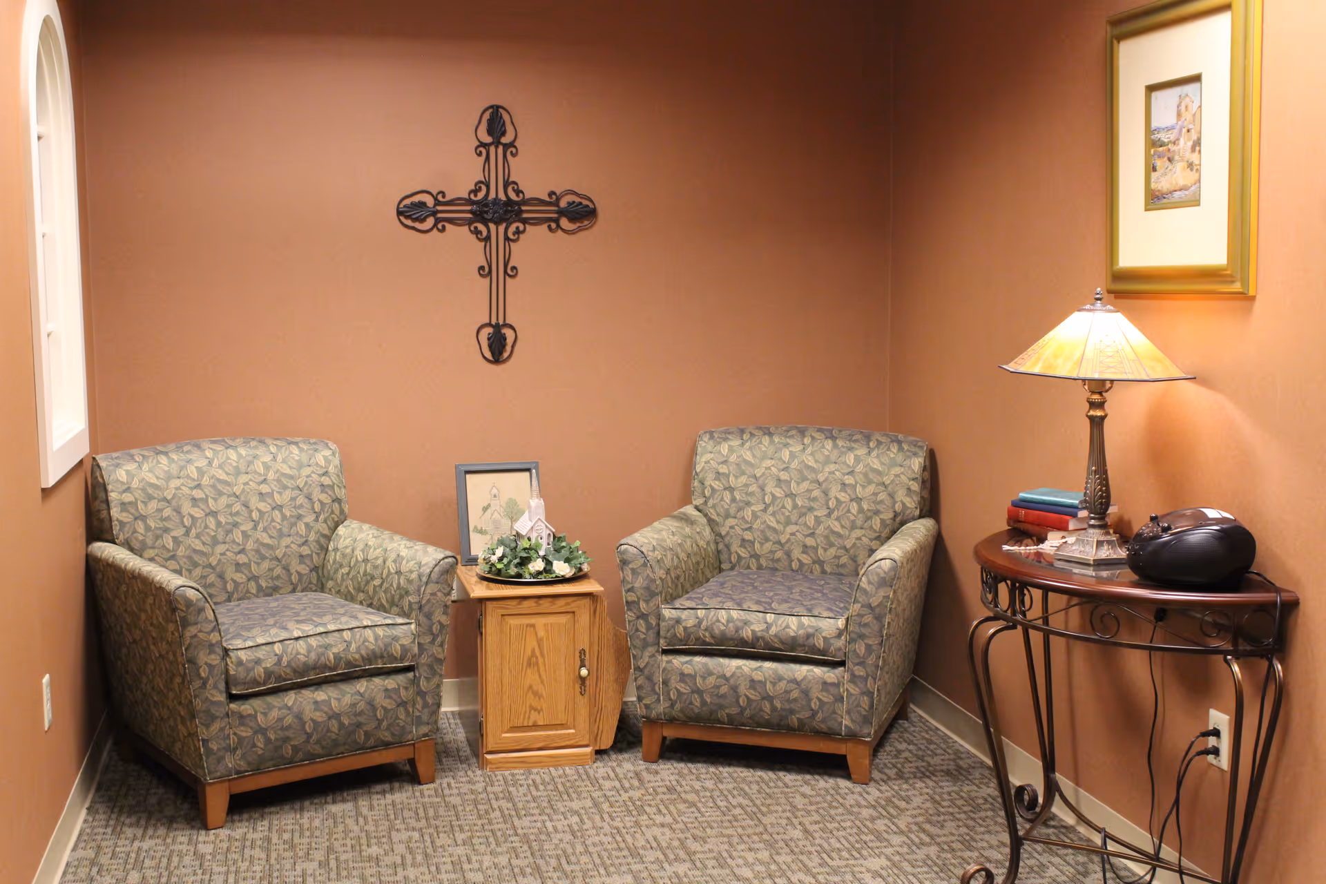 A small sitting area with two patterned armchairs separated by a wooden side table with a floral arrangement and a framed picture. On the right side, there is a decorative metal console table with a lamp, some books, and a black radio or speaker. The walls are painted brown, and there is a decorative metal cross hanging on the wall above the side table.