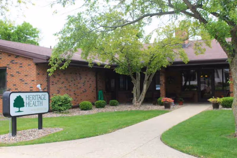 Front entrance of a single-story brick building with a 'Heritage Health' sign, walkway, and trees.