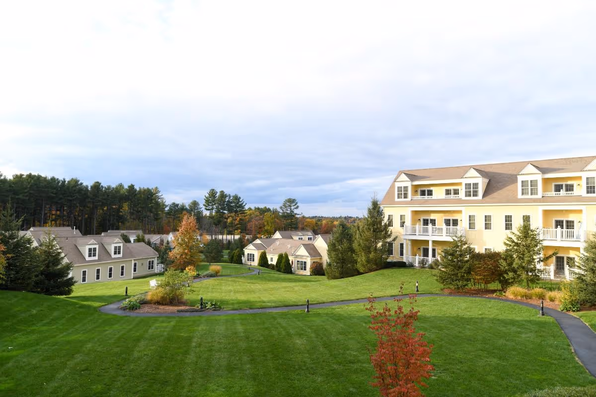 A scenic view of The Commons in Lincoln senior living facility showing well-maintained green lawns, paved walking paths, several residential buildings with beige siding and white trim, surrounded by trees with some autumn foliage under a cloudy sky.