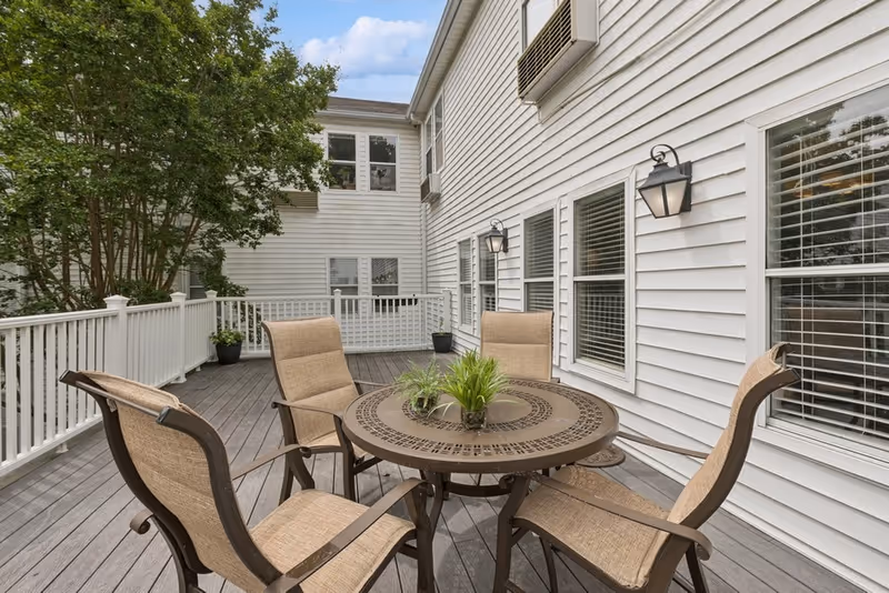 Outdoor patio area with a round metal table and four cushioned chairs on a wooden deck attached to a white building with multiple windows and wall-mounted lantern lights. There are potted plants on the table and deck, and a large tree is visible in the background under a partly cloudy sky.