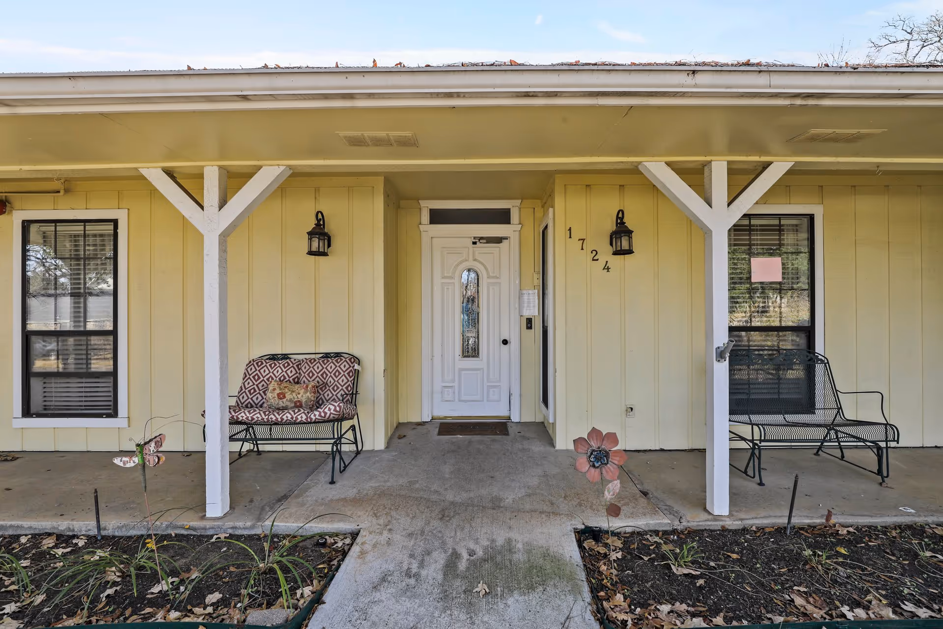 Front porch of a yellow senior living facility unit with a white door numbered 1724, two black-framed windows, two black metal benches with cushions, and decorative garden stakes shaped like a butterfly and a flower in the garden beds.