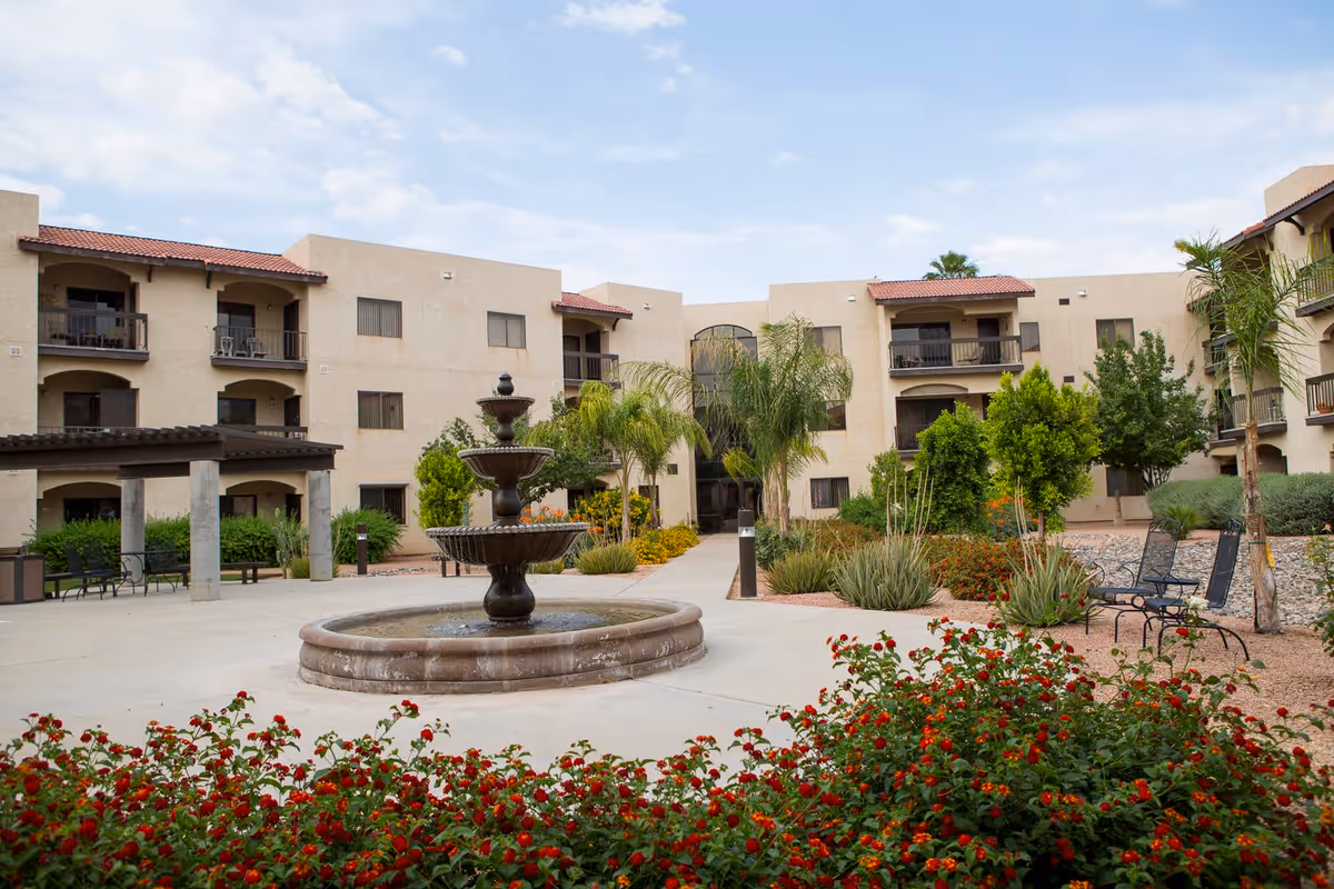 Outdoor courtyard area of a senior living facility with a three-tiered water fountain in the center, surrounded by landscaped plants, flowers, palm trees, and benches. The beige multi-story building with balconies and red-tiled roofs encloses the courtyard under a partly cloudy sky.
