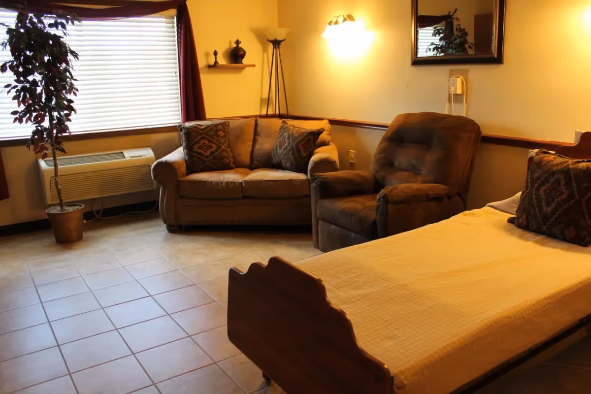 A cozy senior living bedroom with a bed in the foreground, a recliner and loveseat by a window and a potted plant.