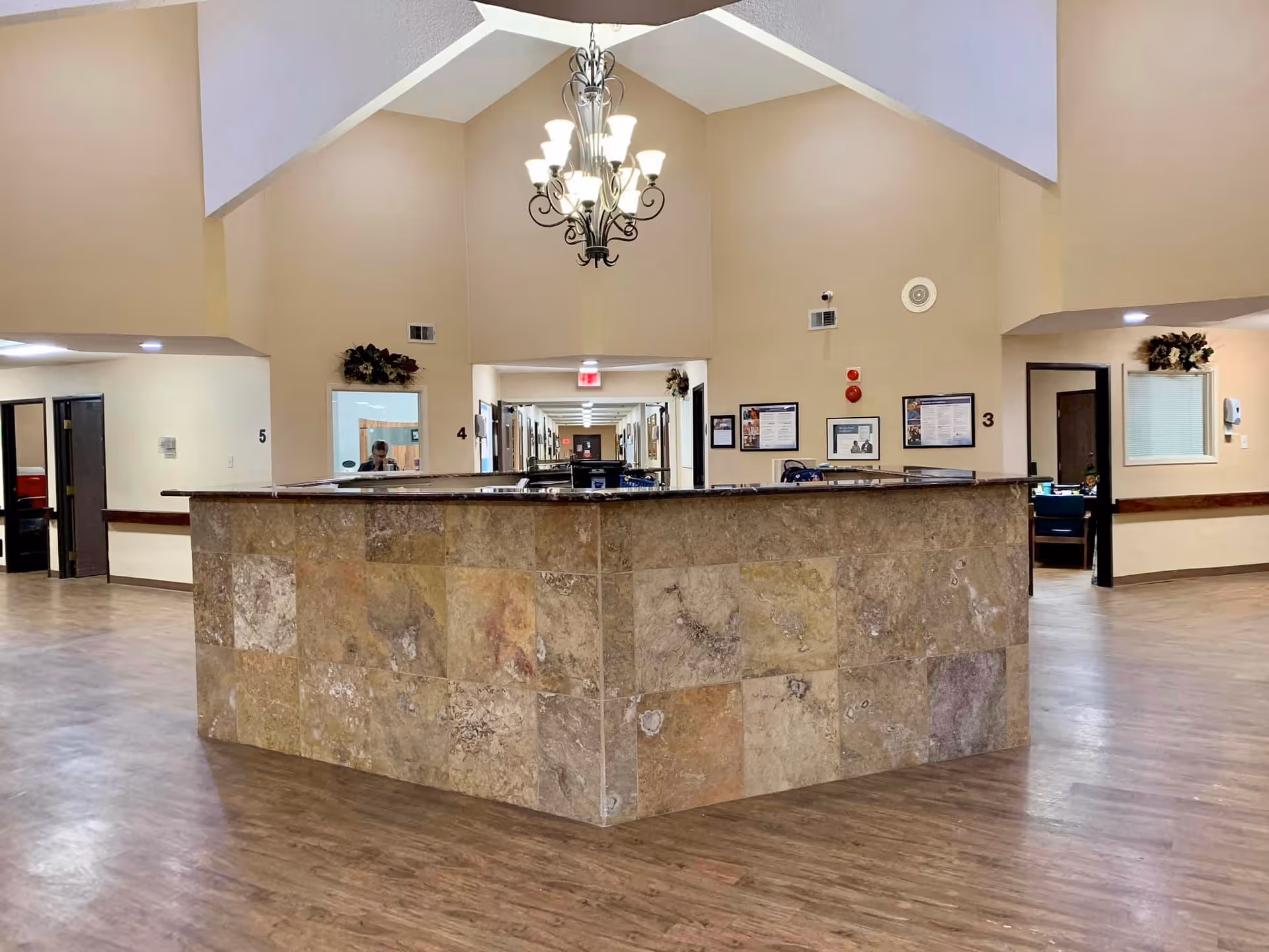 Interior view of a nursing facility reception area with a large stone-tiled reception desk in the center. The space has beige walls, wood flooring, and a chandelier hanging from a vaulted ceiling. Doors and hallways lead to other rooms, and there are framed notices and decorations on the walls.