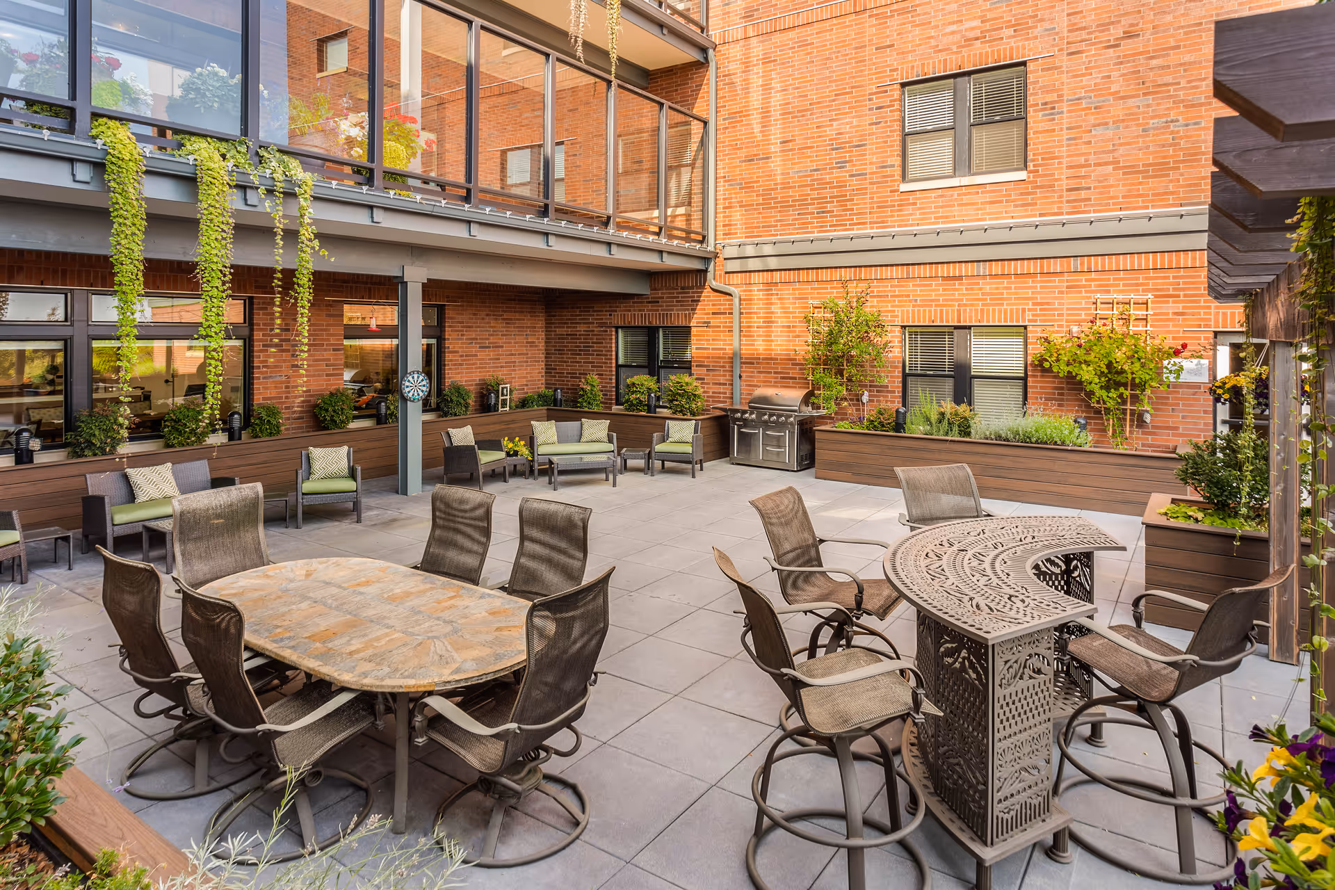 An enclosed sunny courtyard patio with multiple dining tables and chairs, raised planters, hanging greenery, a grill, and brick building walls.