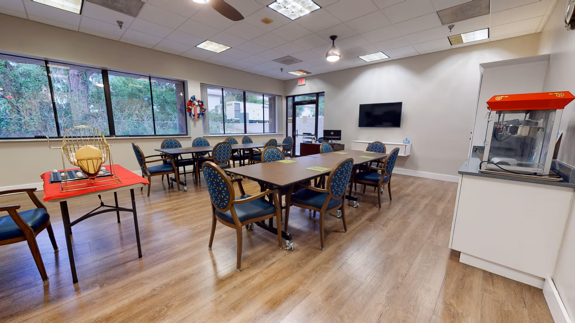 A bright and spacious activity room with multiple tables and chairs arranged for group seating. The room has large windows letting in natural light, a ceiling fan, a wall-mounted TV, and a popcorn machine on a counter. There is also a bingo cage on a small table covered with a red cloth.