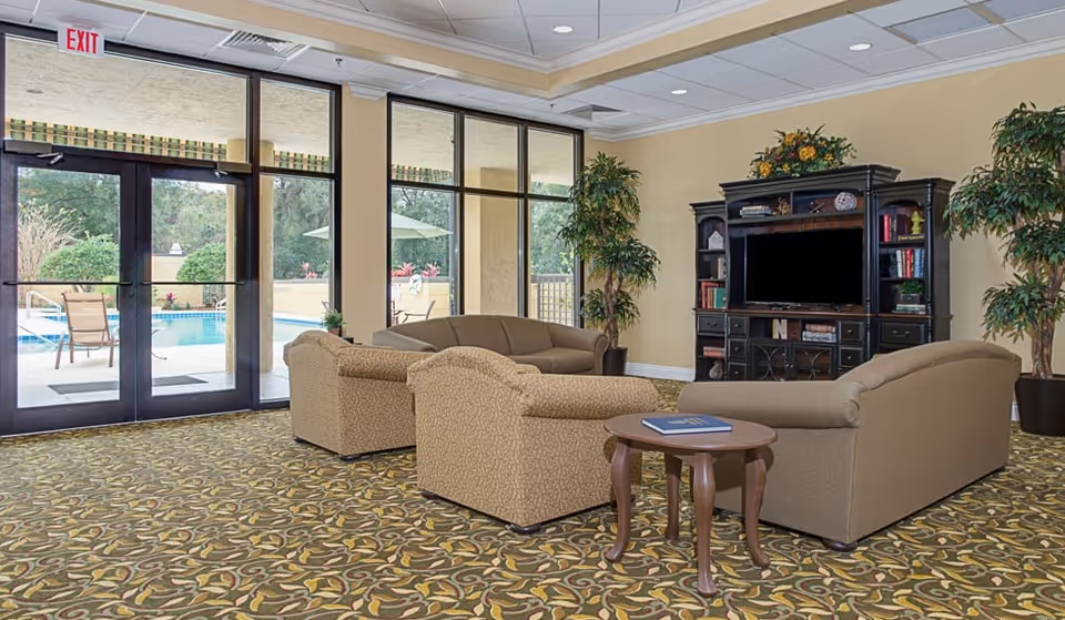 A spacious living room area with patterned carpet, three beige upholstered sofas arranged around a small wooden side table with a book on it, and a large dark wooden entertainment center with a flat-screen TV. Large windows and glass doors provide a view of an outdoor pool area with chairs and umbrellas. Two large potted plants flank the entertainment center.