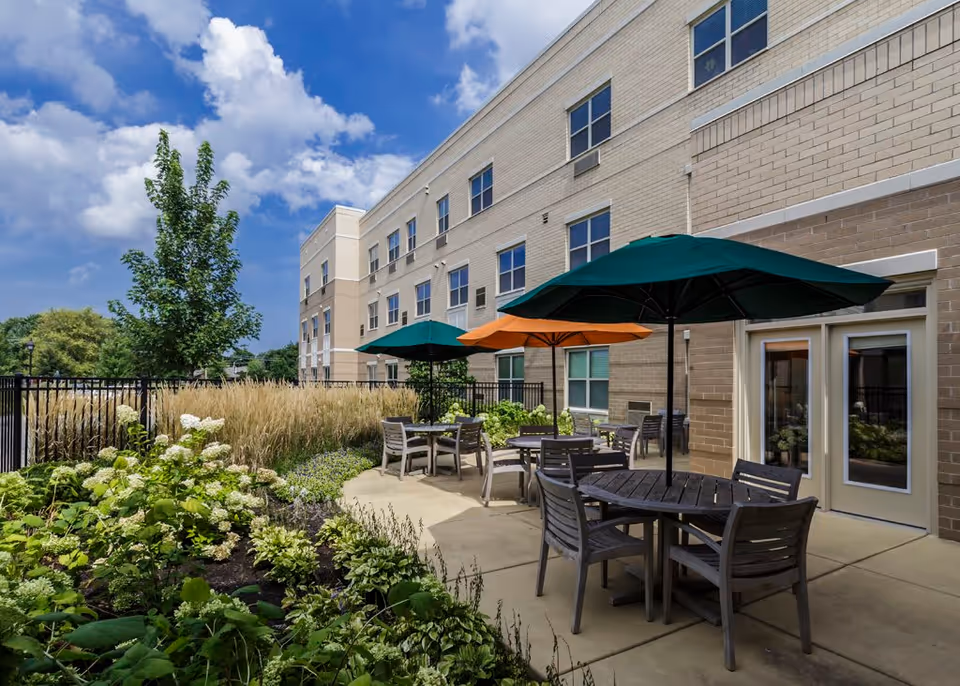 Outdoor patio area at Mercy Circle Senior Living with several round tables and chairs under green and orange umbrellas, adjacent to a multi-story beige brick building. The patio is surrounded by landscaped greenery and plants under a partly cloudy blue sky.