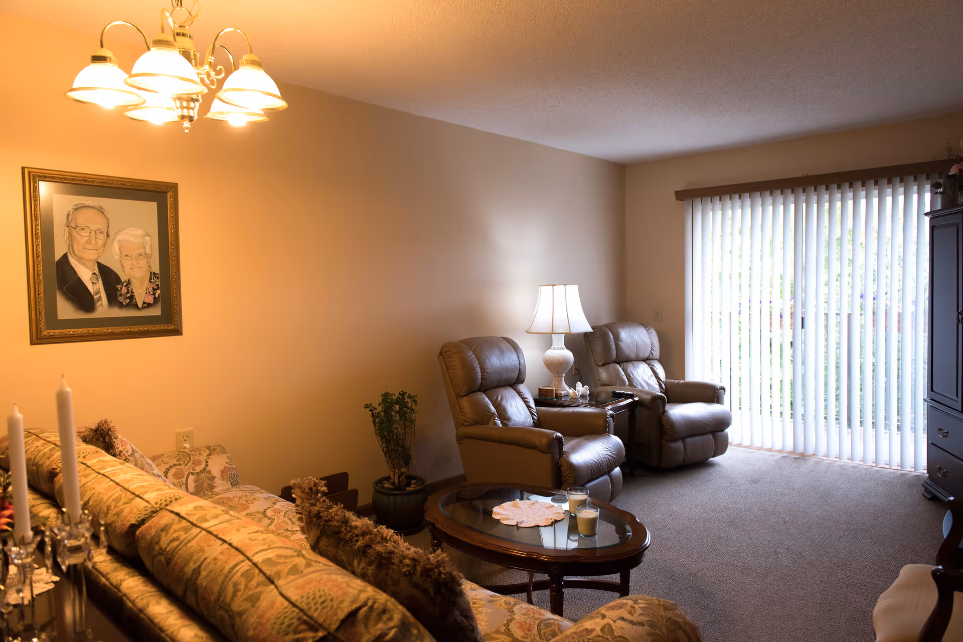 A cozy living room with a floral patterned sofa, two beige recliner chairs, a glass-top coffee table with candles, a side table with a lamp, a framed portrait of an elderly couple on the wall, and vertical blinds covering a sliding glass door.