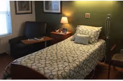 A single bed with patterned bedding and multiple pillows in a cozy room. Next to the bed is a wooden nightstand with a lamp, a phone, and some personal items. There is a dark armchair with a small table beside it near a window with blinds. The walls are painted green and decorated with framed pictures.