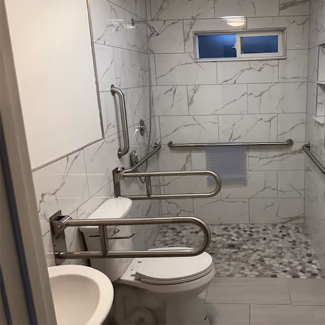 Accessible bathroom featuring a toilet, sink, multiple stainless steel grab bars and a roll-in tiled shower with a pebble floor.