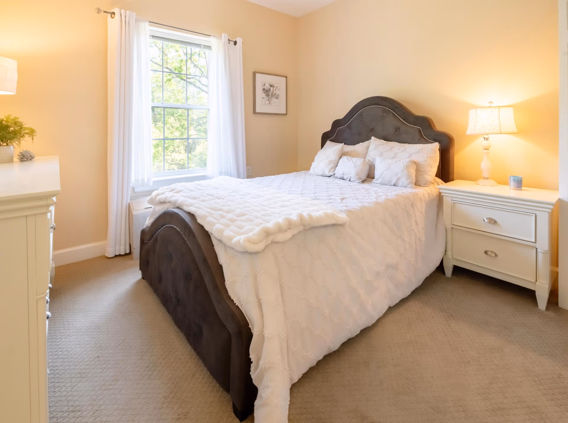 Bright bedroom with a tufted dark headboard, white quilted bedding, nightstands with lamps, and a window with white curtains.