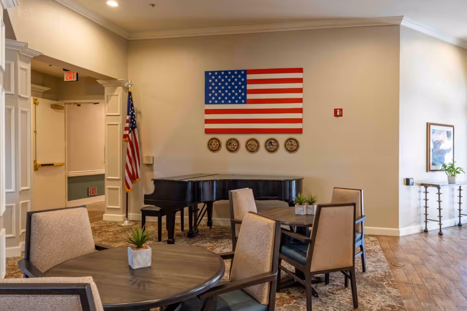 Interior of a senior living facility common area with a grand piano against the wall, an American flag and military insignias displayed above it. There are round tables with chairs around them and small potted plants on the tables. The room has beige walls, carpeted and wooden flooring, and an exit door in the background.