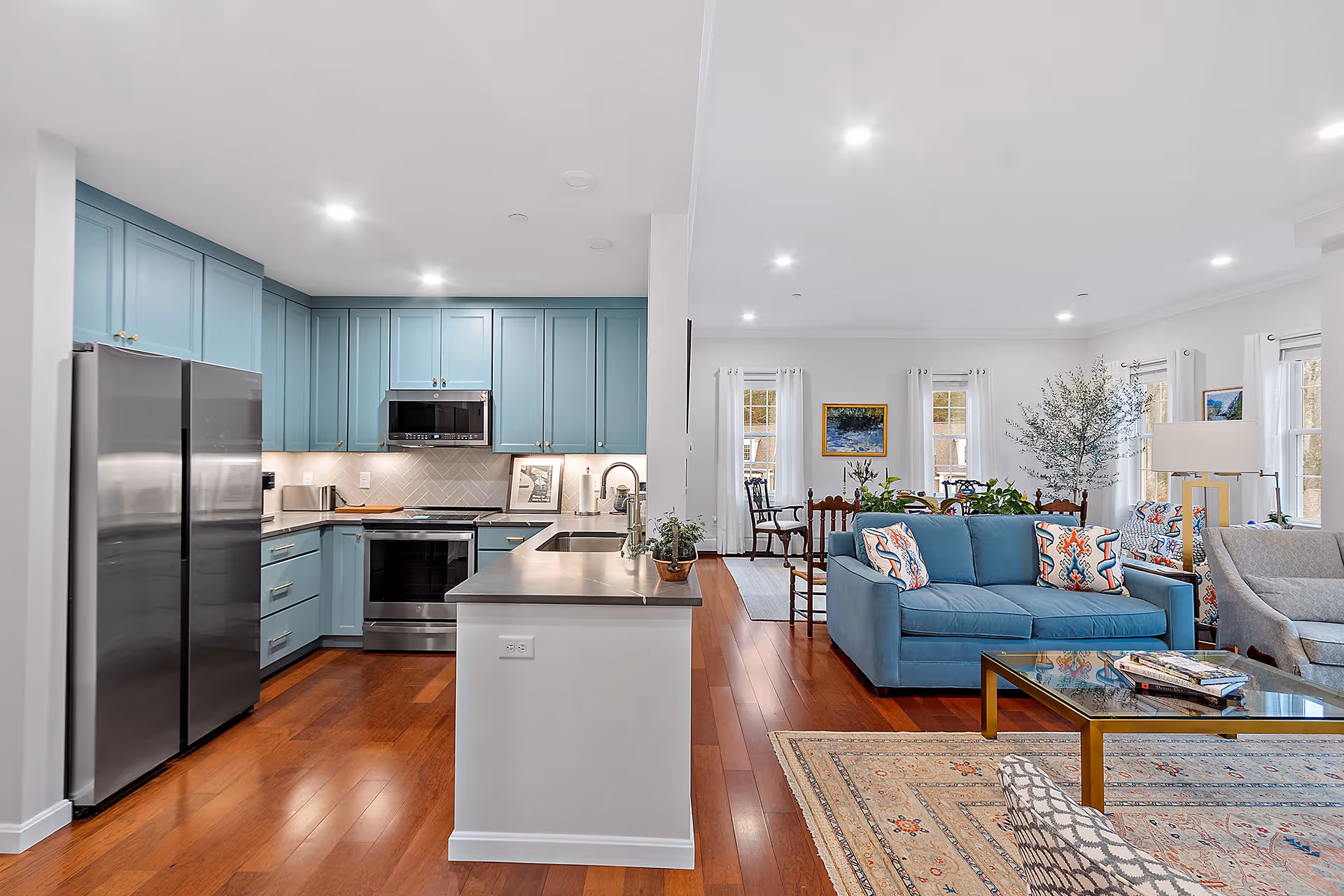 Open-plan interior showing a teal-cabinet kitchen with stainless steel appliances and a connected living area with a blue sofa and hardwood floors.