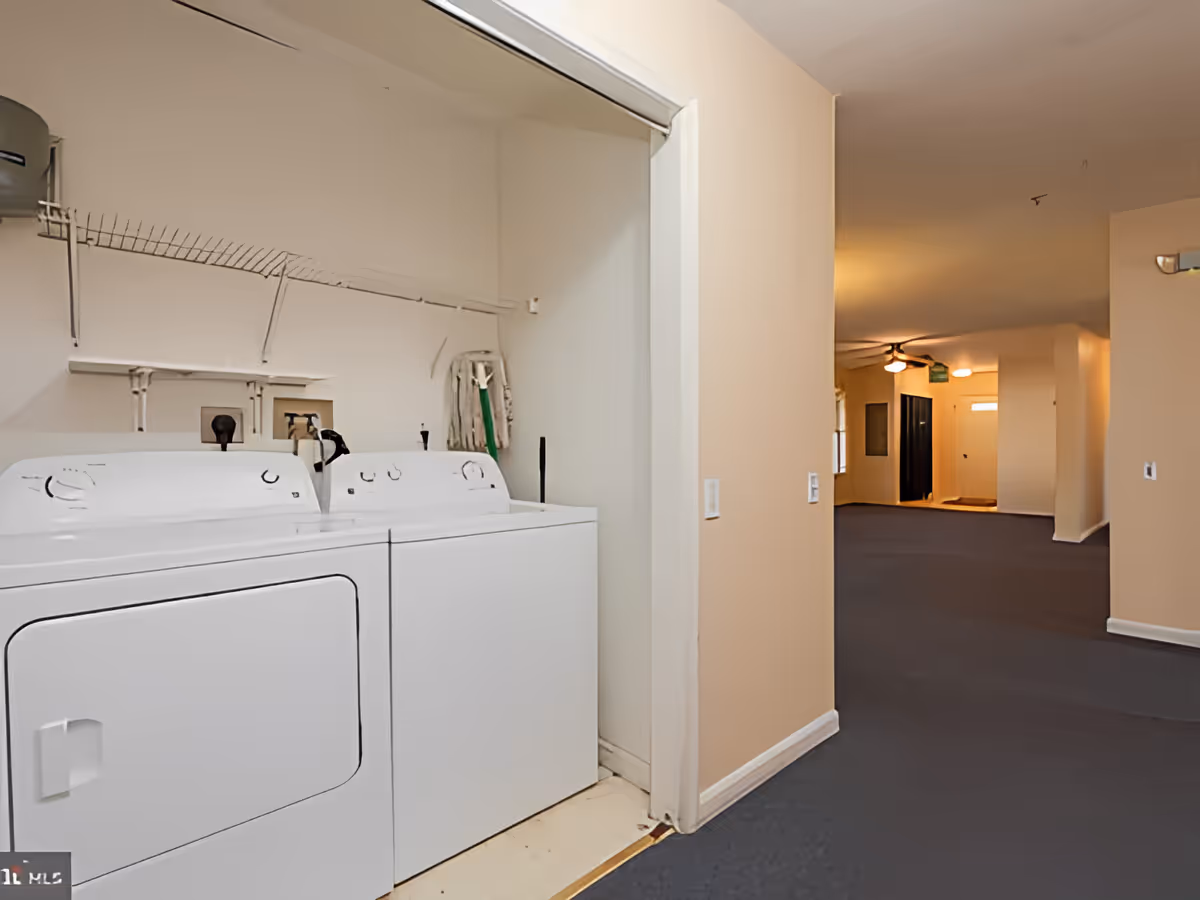 Laundry area with a white washing machine and dryer side by side in a small closet space with a wire shelf above them. The adjacent hallway has beige walls and dark carpet, leading to a ceiling fan and a door in the background.