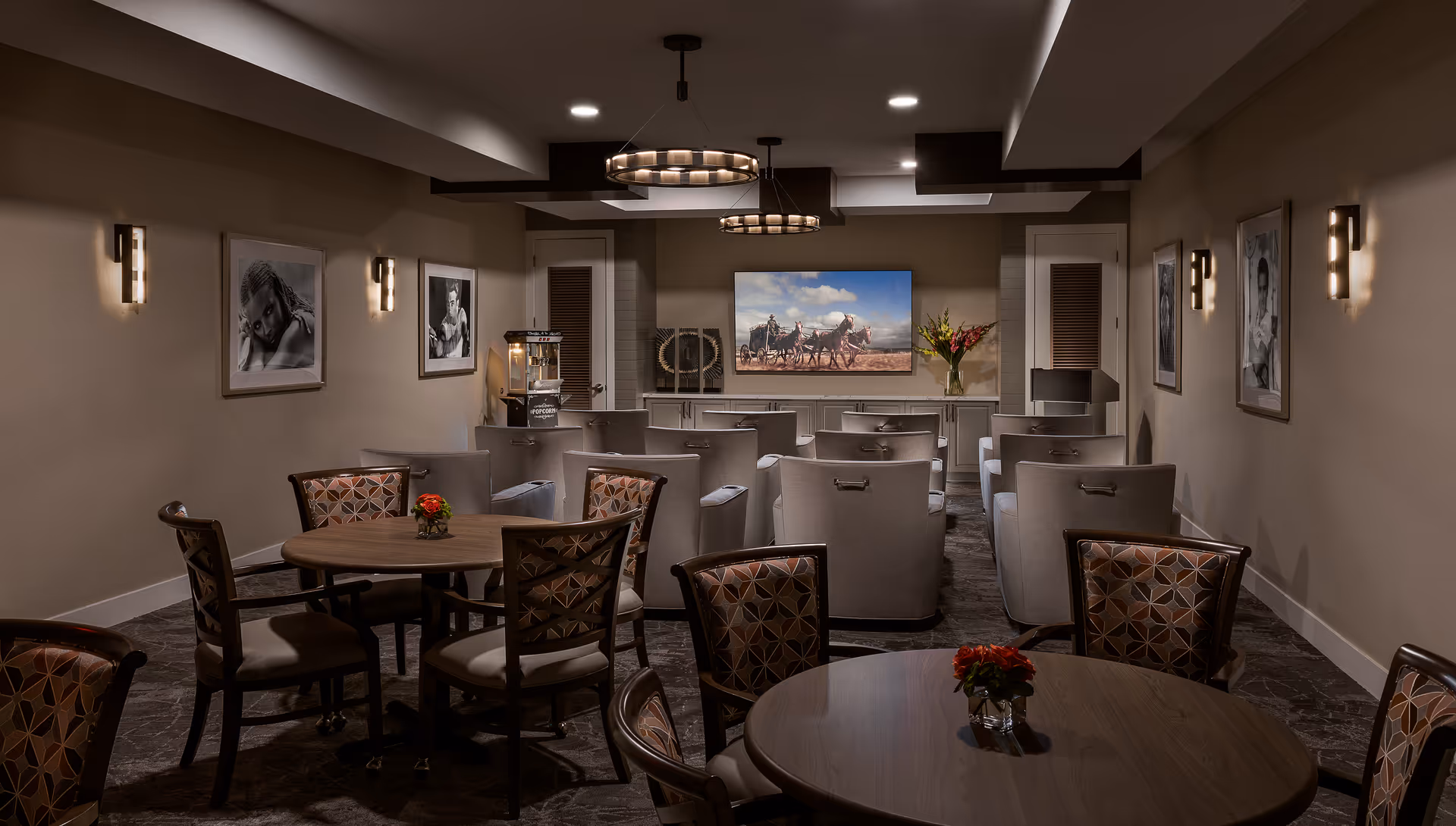 A cozy senior living common room with round tables and patterned chairs in front of rows of upholstered armchairs facing a large wall-mounted TV.