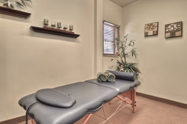 A massage therapy table in a cozy treatment room with folded towels, a potted plant, shelving and wall art.