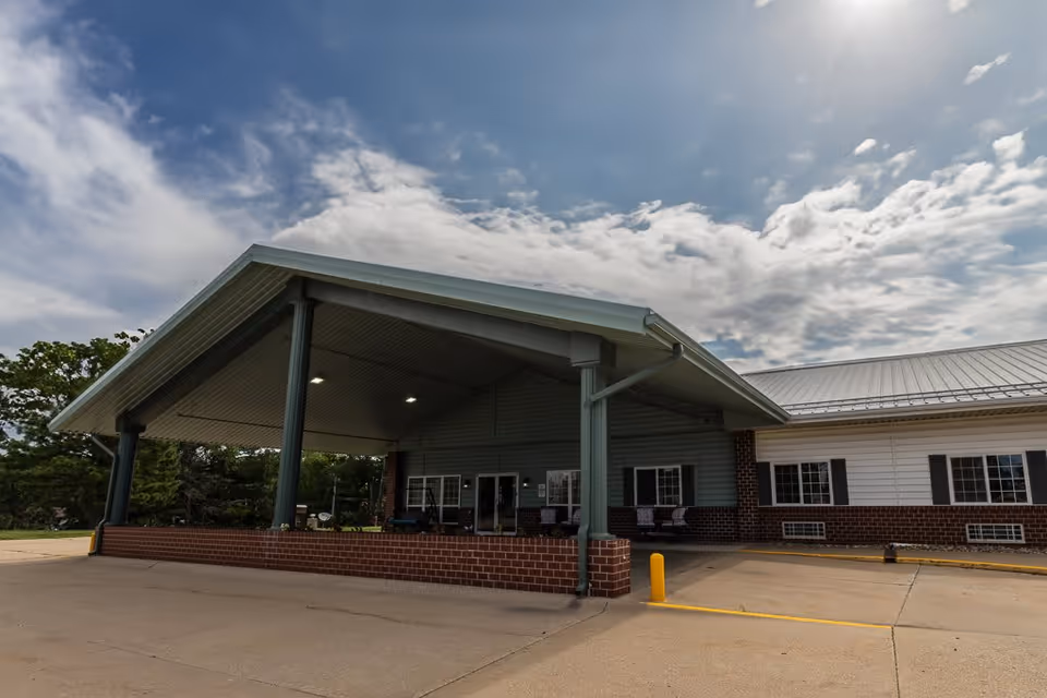 Exterior view of a senior living facility building with a large covered entrance supported by green pillars and a brick base. The building has white siding with brick accents and multiple windows. The sky is partly cloudy.