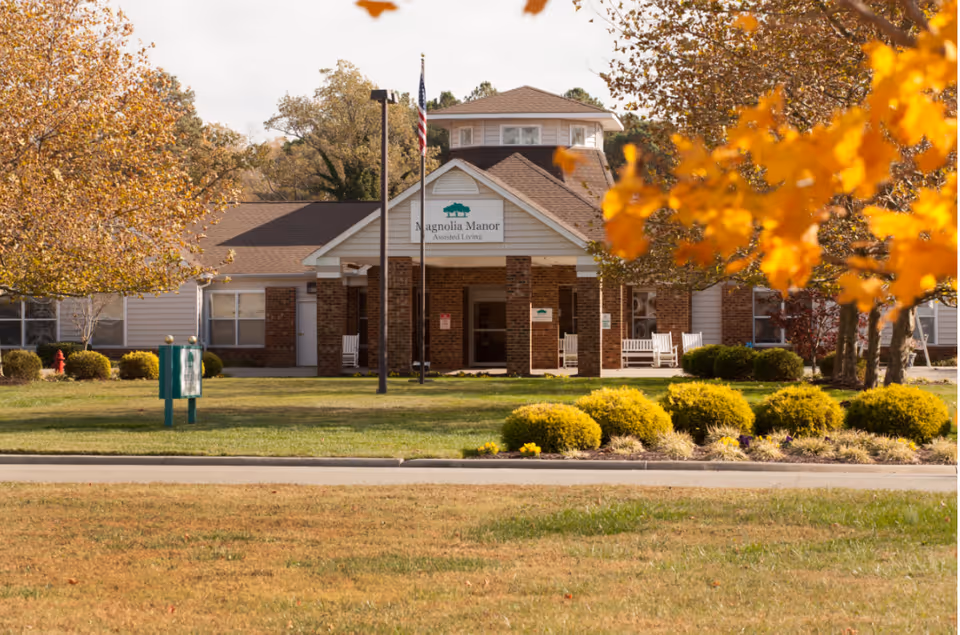 Front exterior view of a senior living facility named Magnolia Manor Assisted Living, surrounded by green grass, bushes, and trees with autumn foliage. The building has a brick and siding facade with a covered entrance and an American flag on a pole in front.