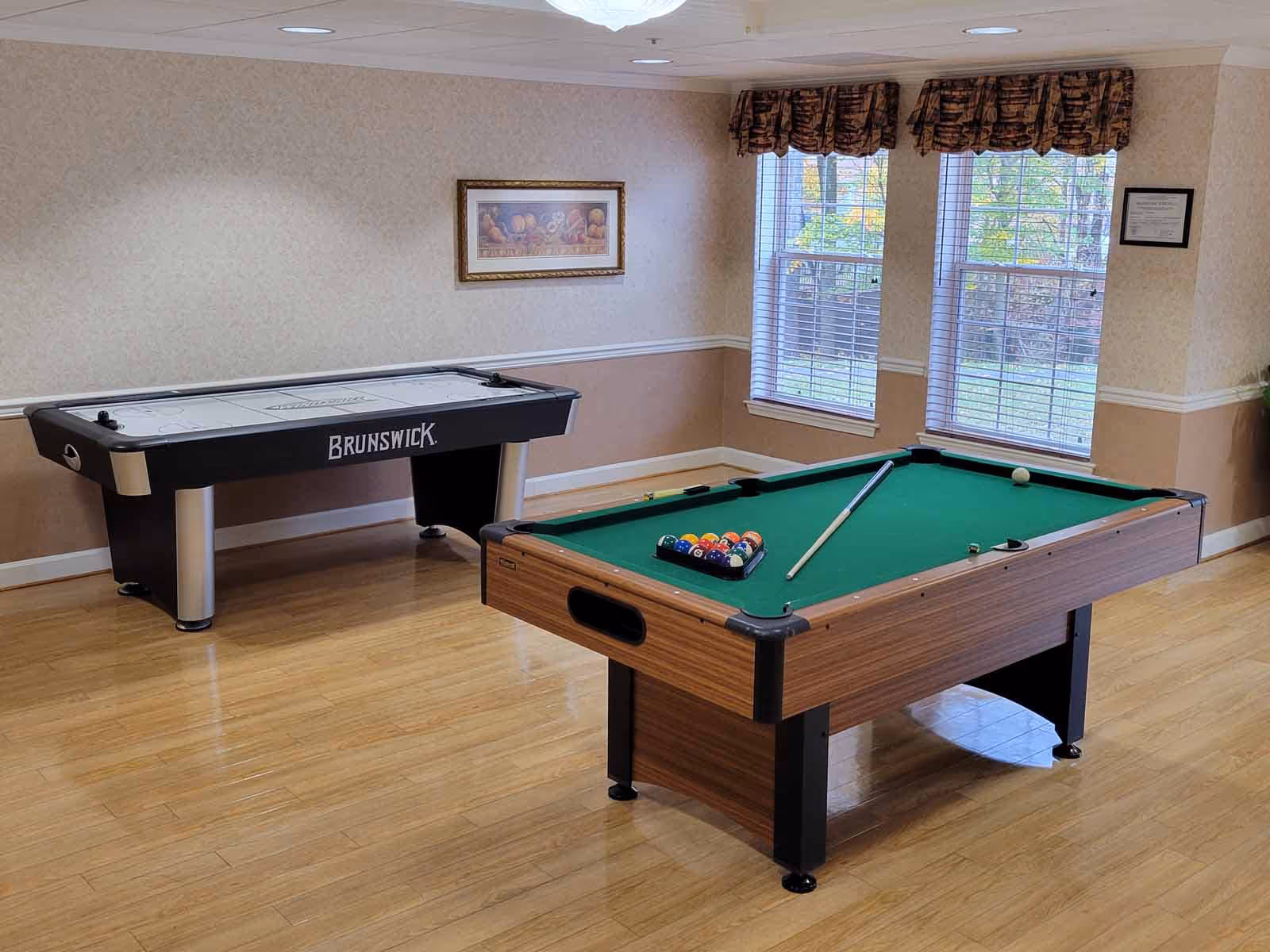 A bright recreation room with a pool table and an air hockey table on a wood floor near windows with blinds.