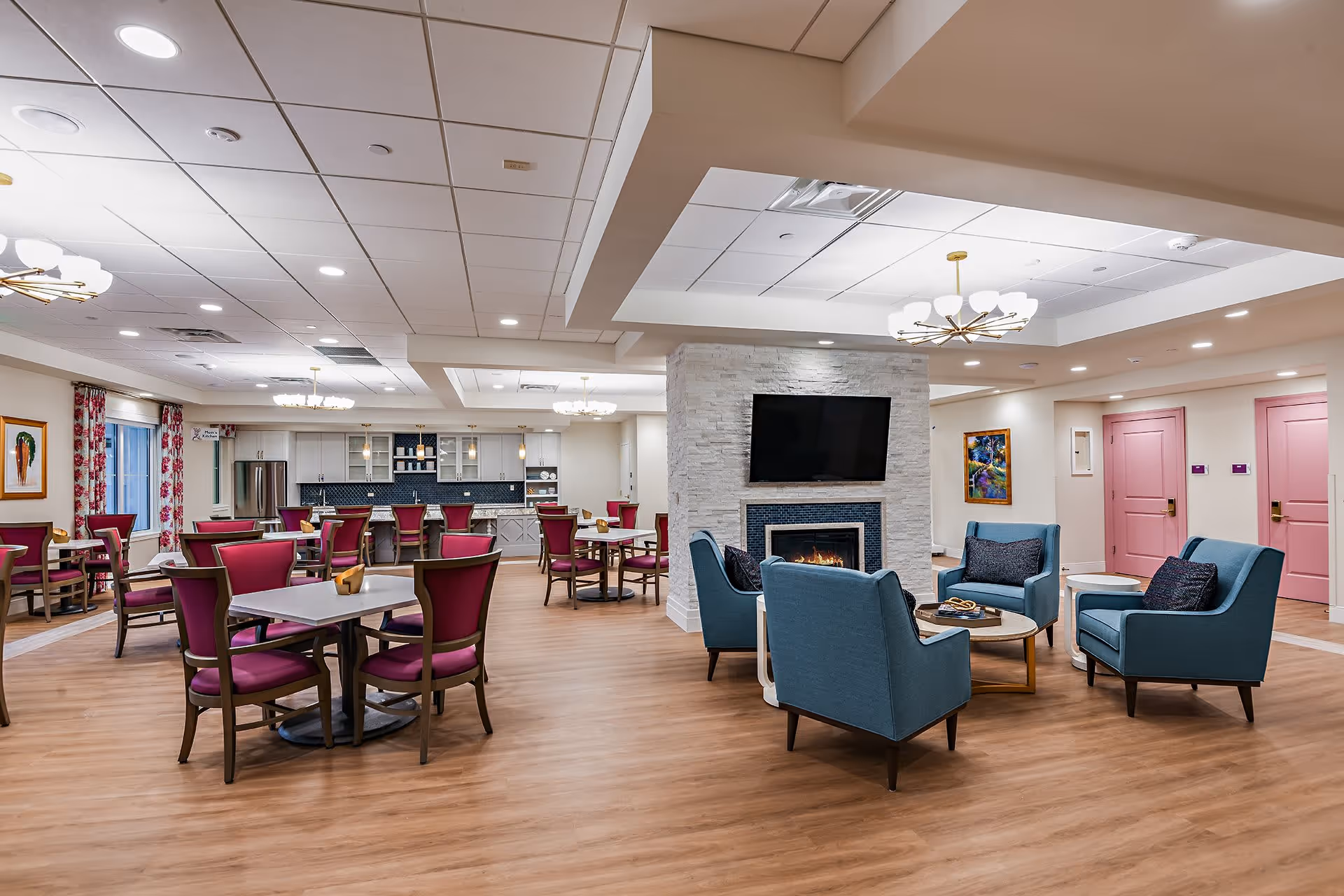 Open communal dining and lounge area with pink dining chairs, blue armchairs around a central fireplace, and a kitchen counter in the background.