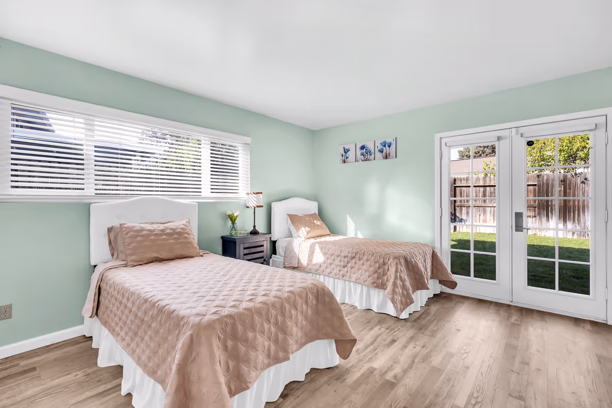 A bright bedroom with two single beds covered in light brown quilted bedspreads and white bed skirts. The room has light green walls, a long horizontal window with white blinds, a small black nightstand with a lamp and a vase of flowers between the beds, and a set of French doors leading to a grassy outdoor area enclosed by a wooden fence. Three small floral paintings hang on the wall above one of the beds.
