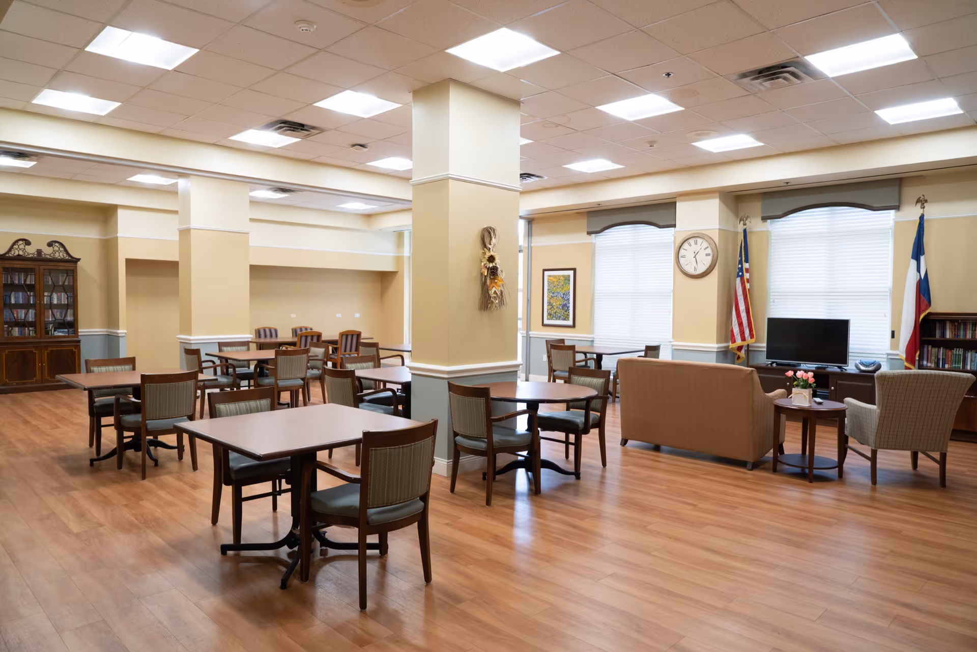 A spacious common area in a senior living facility with multiple wooden tables and chairs arranged for dining or socializing. The room has light-colored walls with white trim, a wooden floor, and several columns. There is a seating area with a sofa, armchairs, a TV, and two flags near the windows. A clock and framed artwork are mounted on the walls.