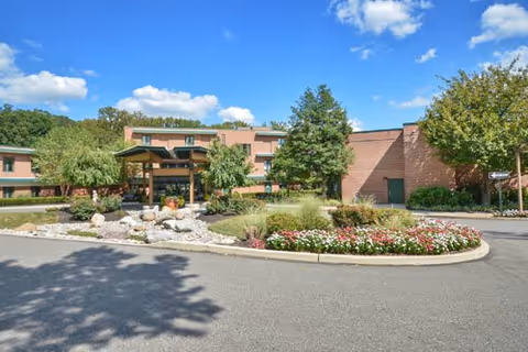 Exterior view of Wesley Enhanced Senior Living facility showing a circular driveway with landscaped flower beds and trees under a blue sky with scattered clouds.