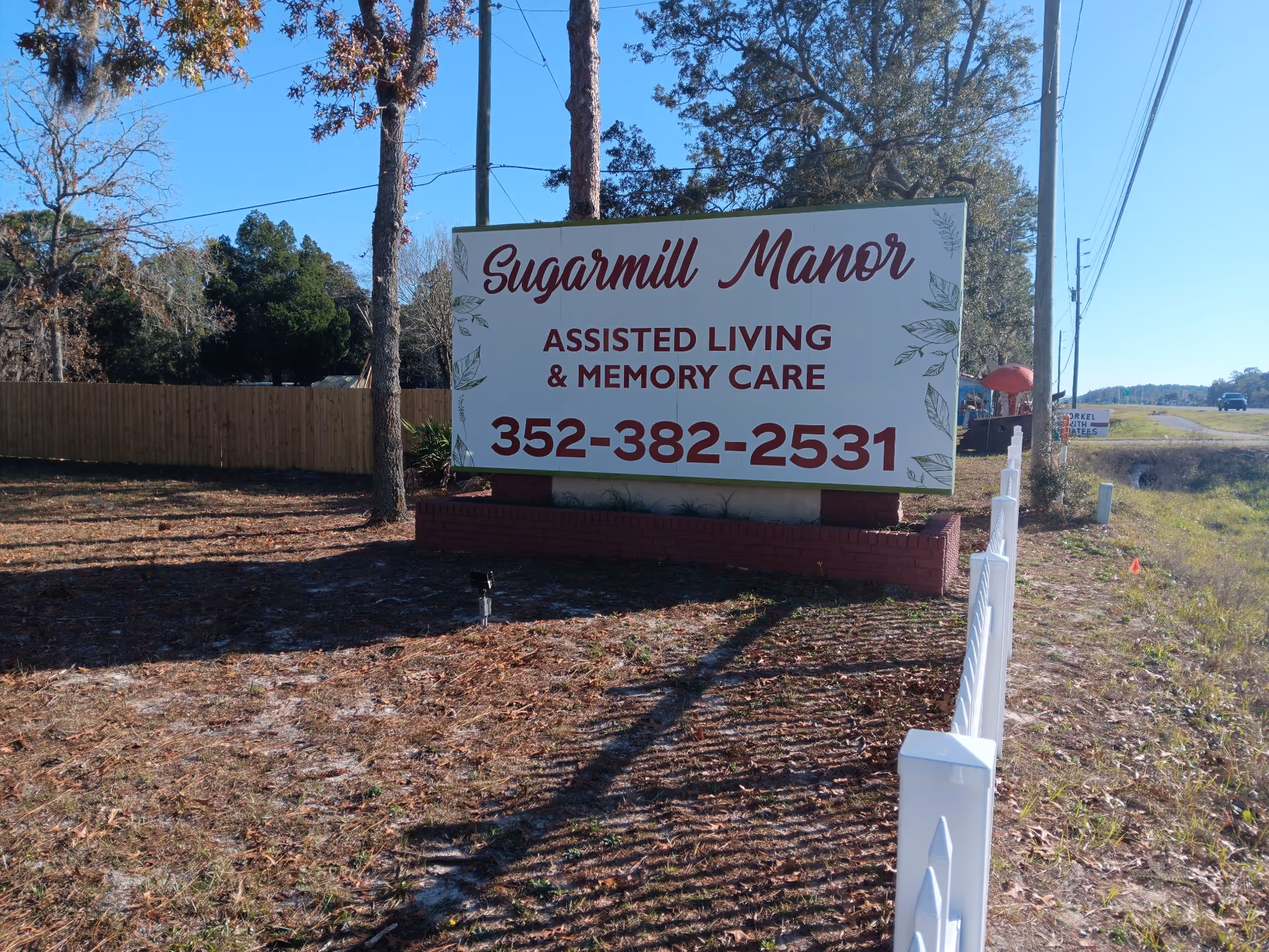 Outdoor image showing a large sign for Sugarmill Manor Assisted Living & Memory Care with a phone number 352-382-2531. The sign is mounted on a brick base next to a white fence, with trees and a road visible in the background under a clear blue sky.