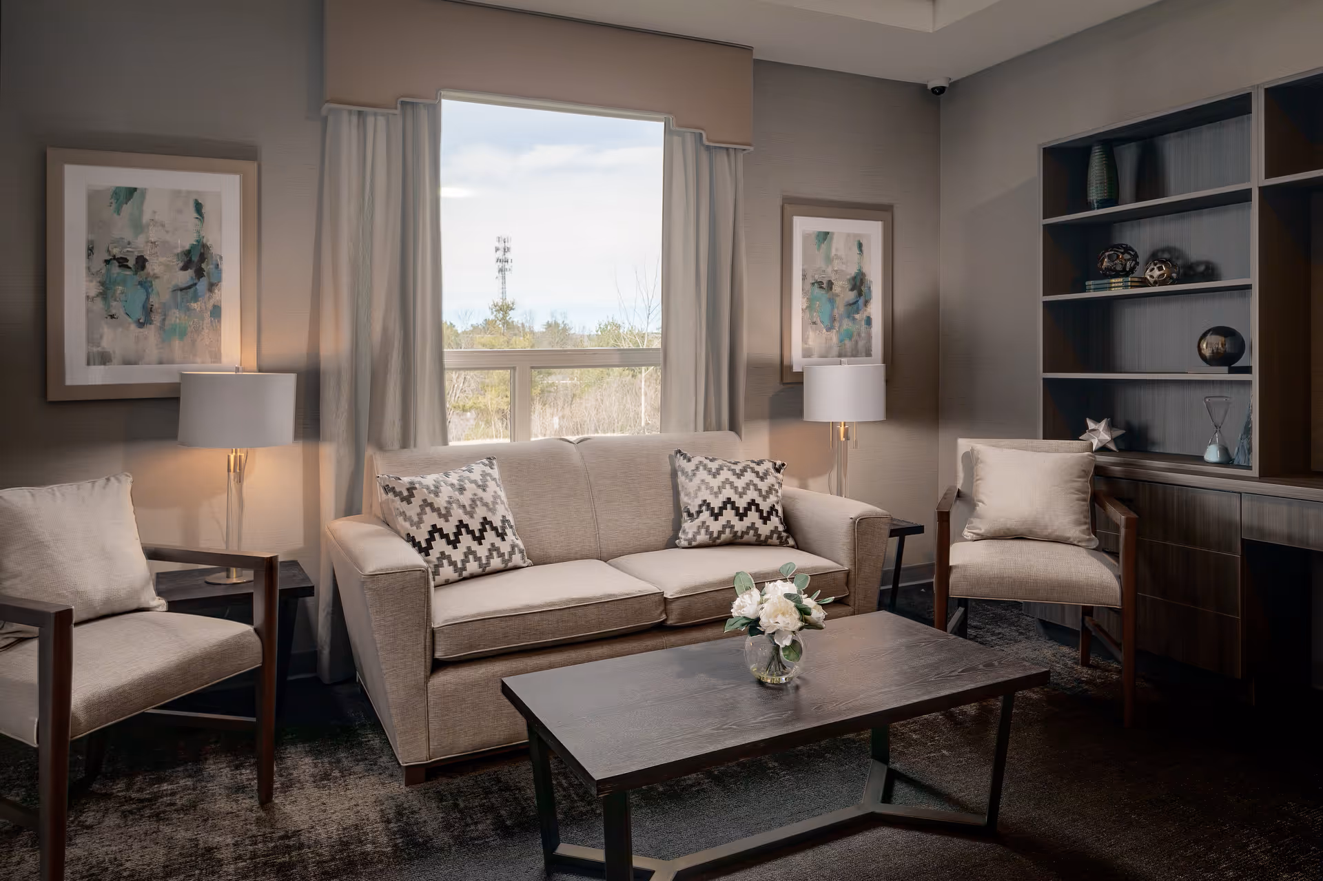 A cozy living room with a sofa, two armchairs, a coffee table with flowers, lamps, artwork, and built-in shelving by a window.