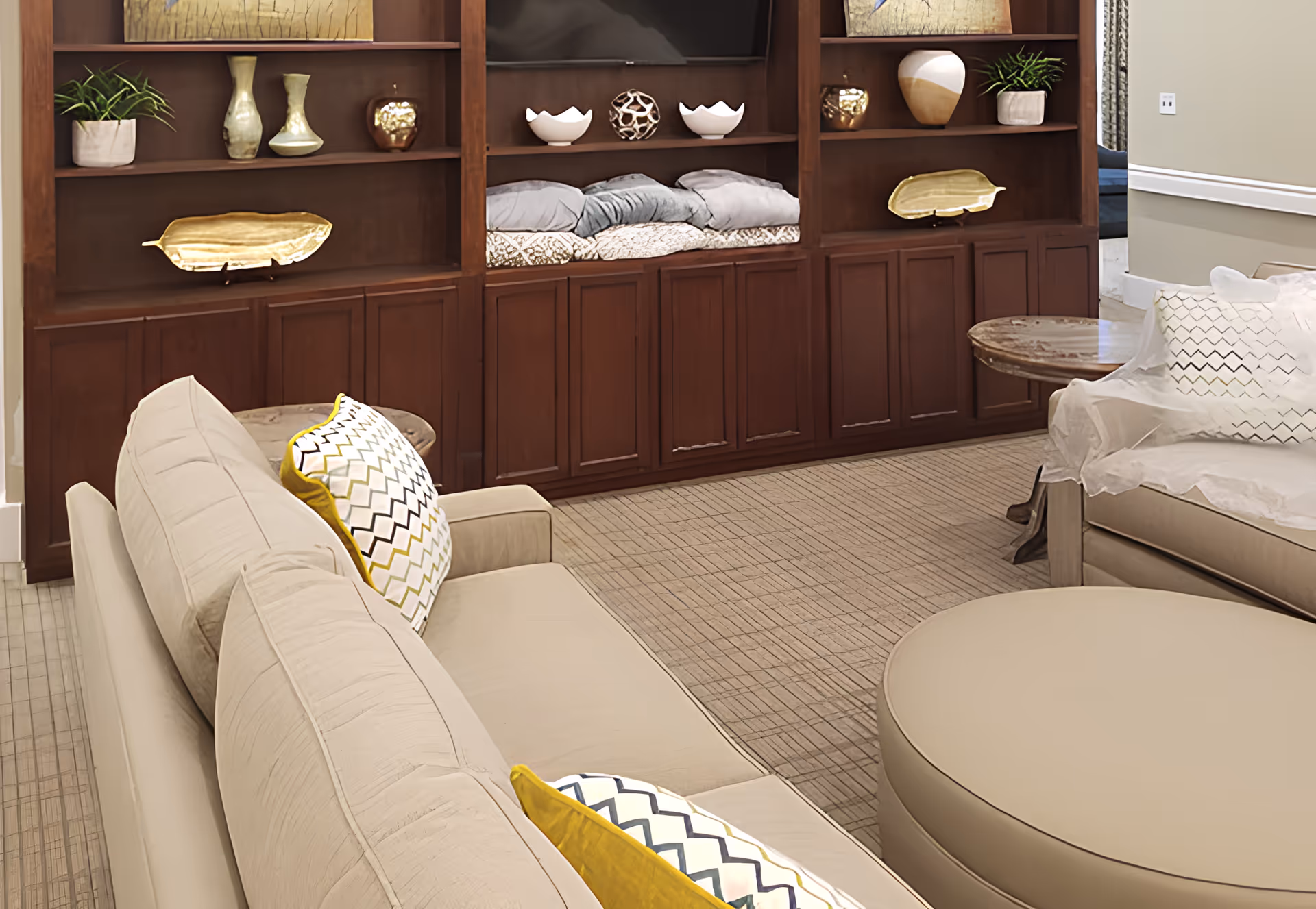 Cozy living room with beige sofas, patterned throw pillows, a round ottoman, and built-in wooden shelving with decorative items.