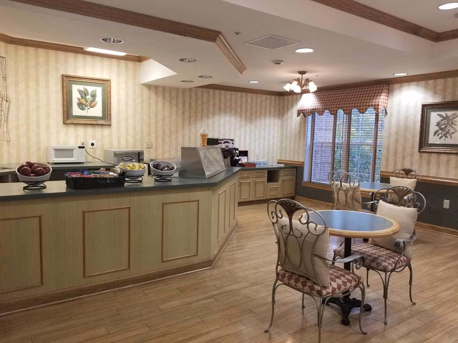 A cozy dining area in a senior living facility with a counter displaying bowls of fruit and snacks, a microwave, and a toaster oven. There are round tables with cushioned chairs, framed botanical artwork on the walls, and a large window with blinds and a valance letting in natural light.