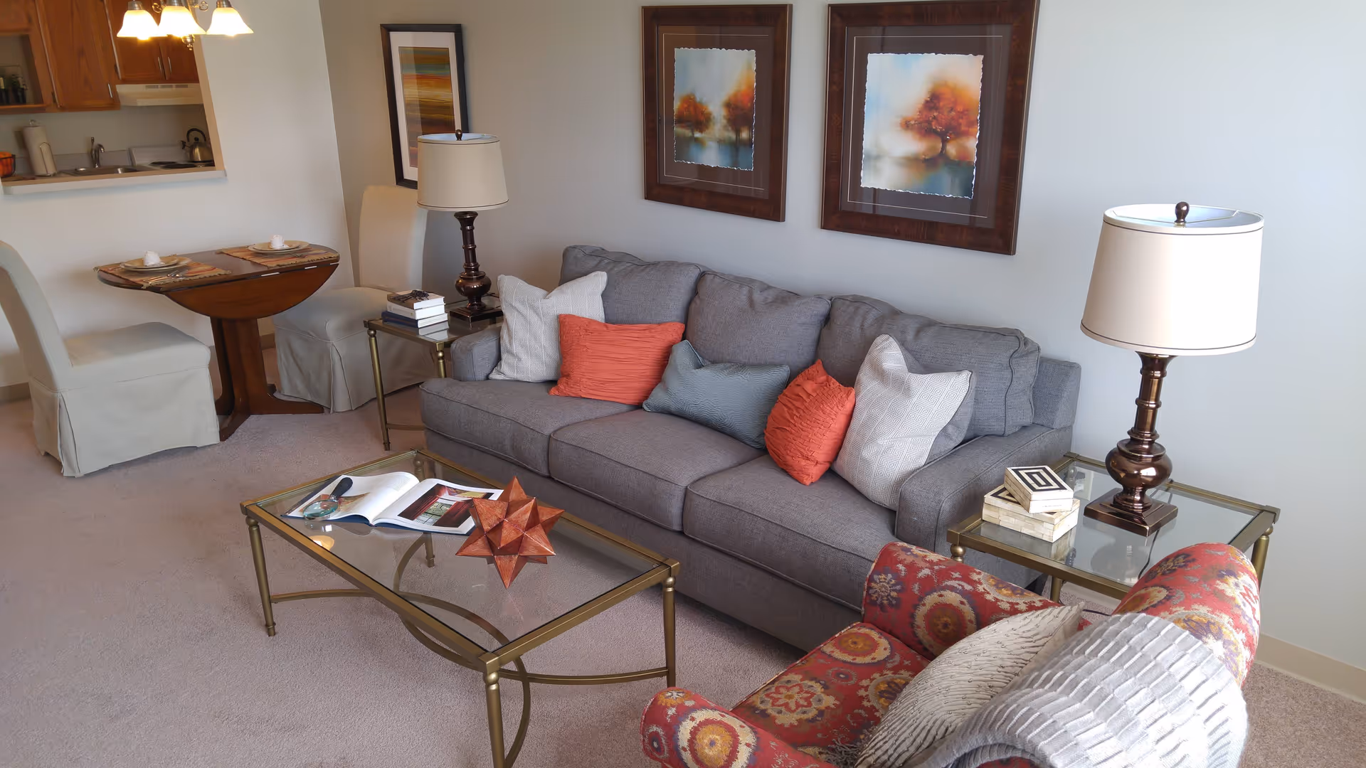 A cozy living room area with a gray sofa adorned with multiple colorful cushions, a glass coffee table with a decorative object and an open book, a patterned armchair with a throw blanket, two side tables each with a lamp, and a small dining table with two chairs near a kitchen pass-through window. Two framed paintings of autumn trees hang on the wall above the sofa.