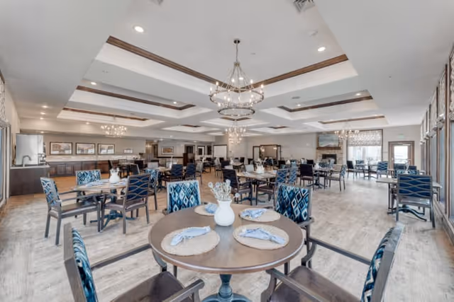 Spacious dining room with round and rectangular tables, patterned chairs, chandeliers, and large windows.