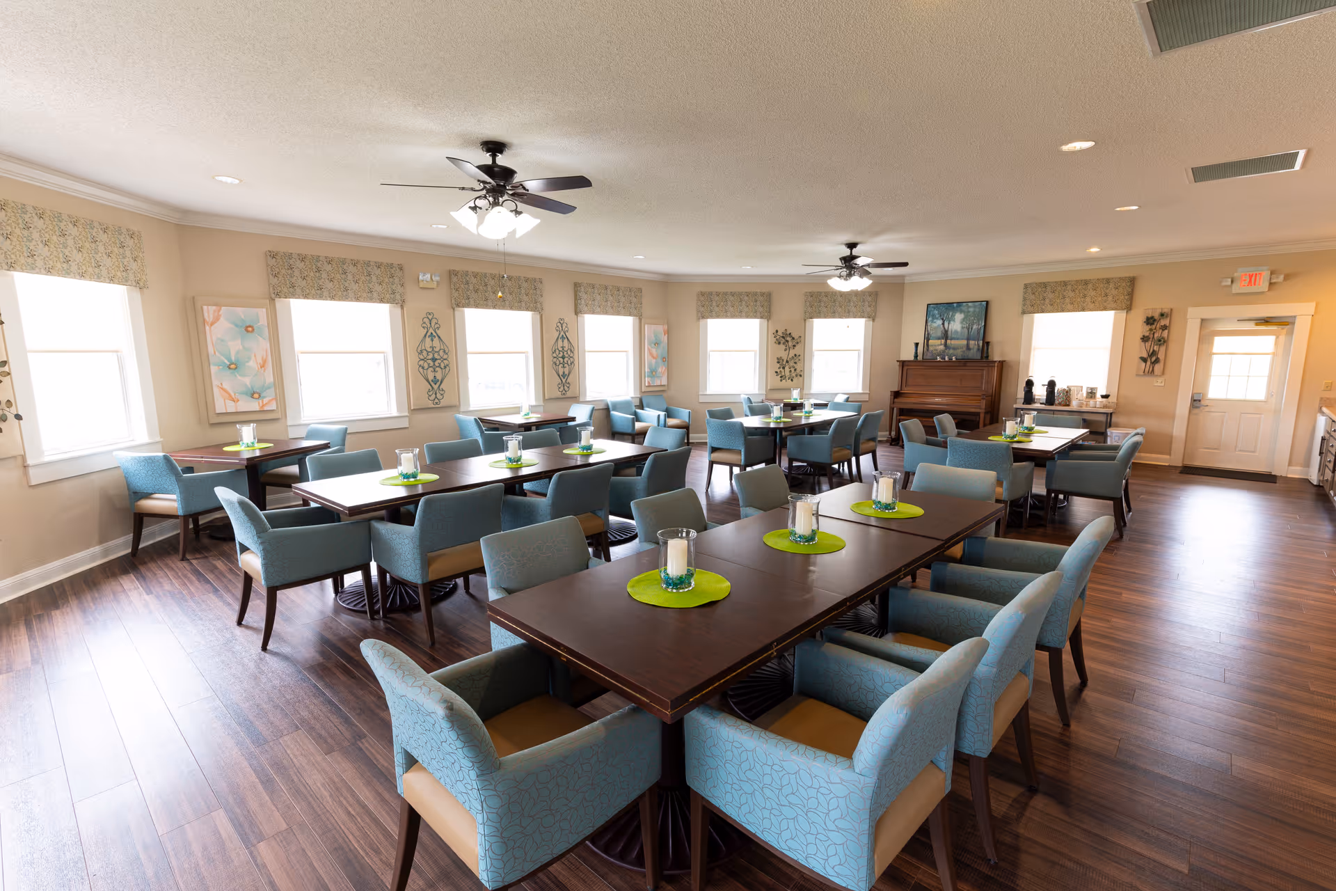 Spacious dining room with multiple dark wood tables surrounded by teal upholstered chairs, green placemats and candle centerpieces, hardwood floors and ceiling fans.