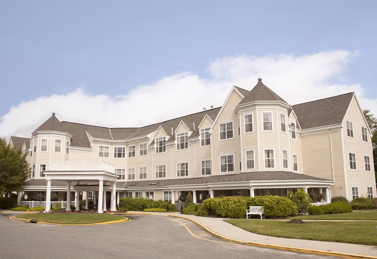 Exterior view of a large, three-story senior living facility building with beige siding, multiple windows, and a covered entrance. The building is surrounded by green bushes, a lawn, and a sidewalk under a partly cloudy sky.