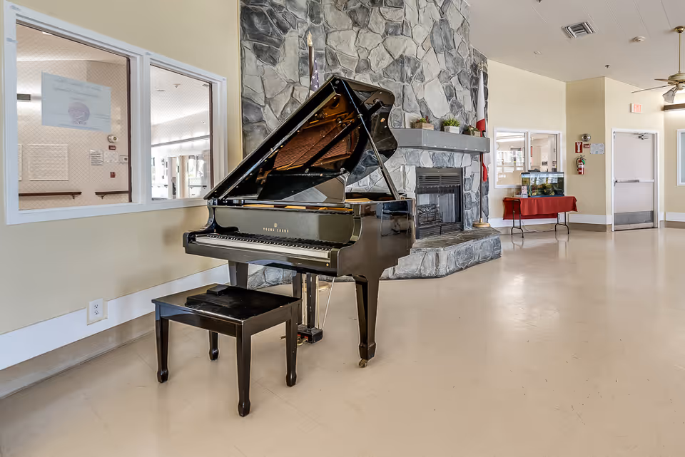 Interior view of a common area in Meadowood Nursing Center featuring a black grand piano with a matching bench in front of a large stone fireplace. The room has beige walls, a ceiling fan, and a table with a red tablecloth holding a fish tank. There are windows and doors visible in the background.