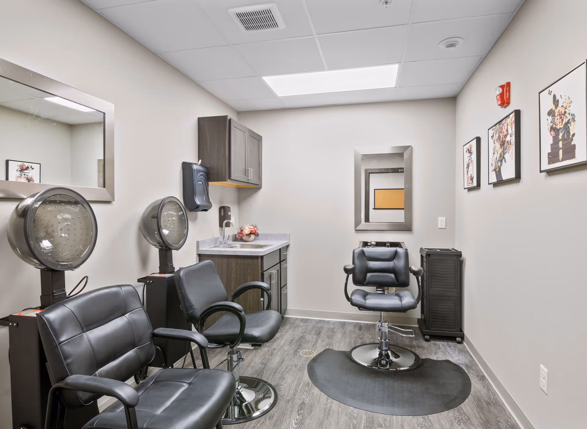 Interior of a small hair salon room with three black salon chairs, two hair dryers, a sink with countertop and cabinets, two mirrors on the walls, and floral artwork on the right wall.