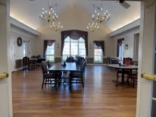 Spacious dining room with wooden floors, multiple dark wood tables and chairs arranged neatly, large windows with curtains allowing natural light, and modern chandeliers hanging from a high ceiling.