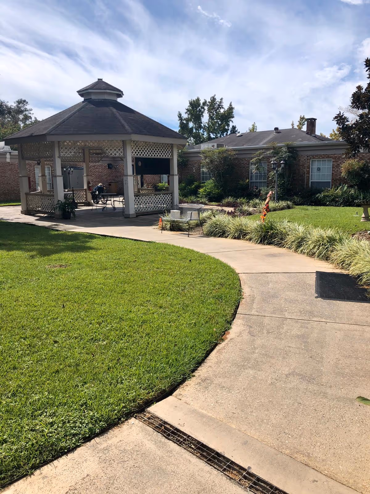 Outdoor view of a senior living facility featuring a paved walkway curving around a well-maintained grassy area leading to a gazebo with lattice sides. The gazebo has a dark shingled roof and contains tables and chairs. In the background, there is a brick building with windows and surrounding greenery under a partly cloudy sky.