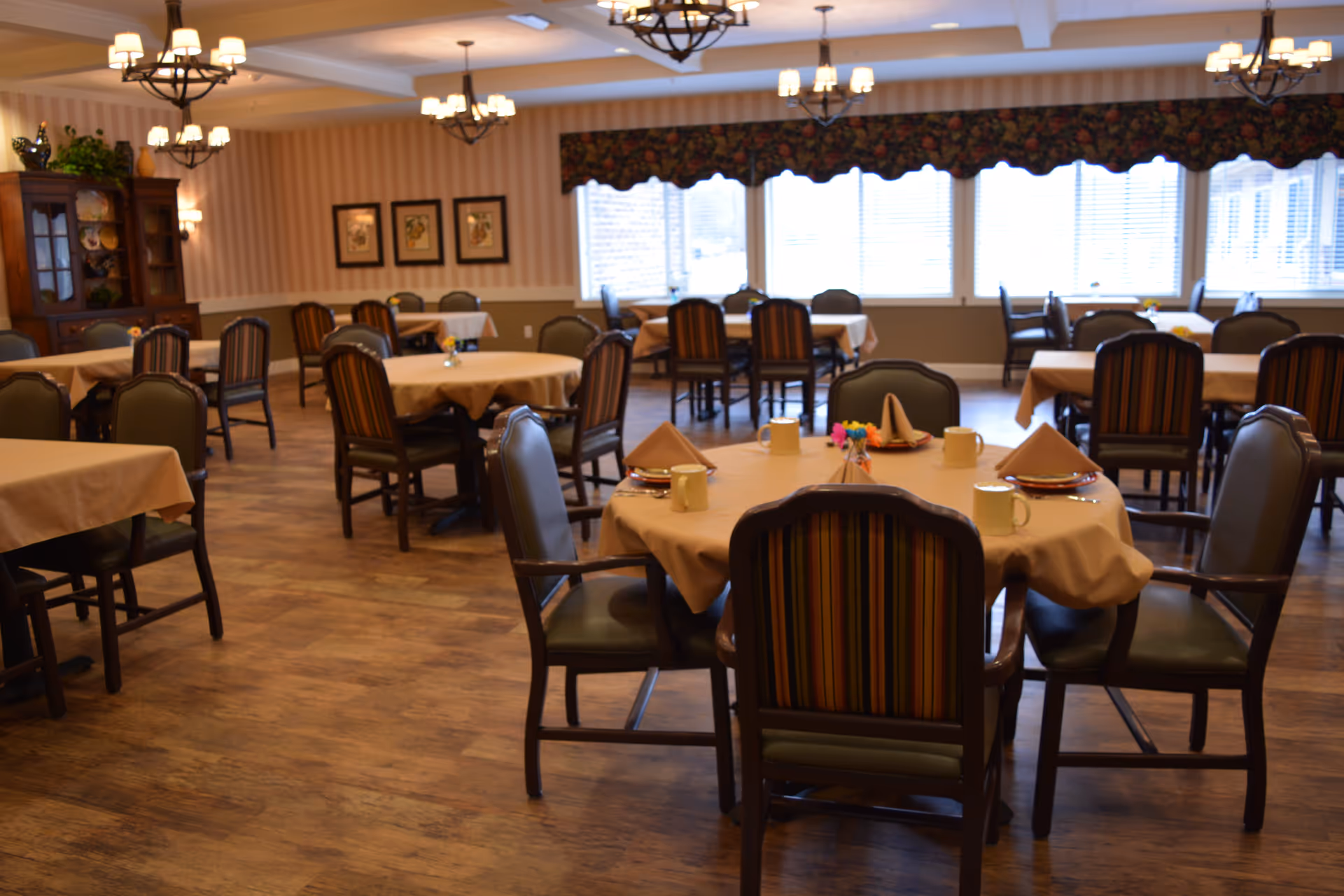 A dining room with multiple round and rectangular tables covered with beige tablecloths. Each table is surrounded by wooden chairs with striped upholstery. The room has wooden flooring, chandeliers hanging from the ceiling, framed artwork on the walls, and large windows with floral valances letting in natural light.