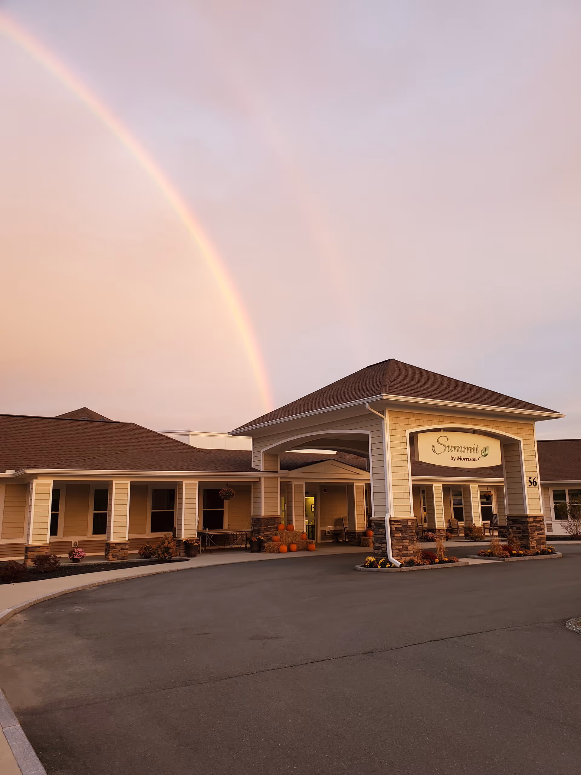 Exterior view of the Summit by Morrison senior living facility at sunset with a double rainbow in the sky. The building features a covered entrance with stone accents and pumpkins arranged near the doorway, along with some landscaping and a paved driveway.
